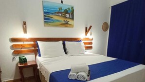 Una cama bien hecha con ropa de cama blanca y azul, un cuadro de una playa en la pared y una cortina azul en la ventana.