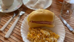 Un desayuno con huevos revueltos, un sándwich y un vaso de zumo de naranja sobre una mesa de madera.