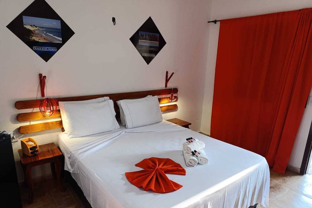 Un lit bien fait, avec du linge de lit blanc, une serviette rouge et des articles de toilette posés sur le lit. Il y a deux tables de chevet avec des lampes, et un rideau rouge à la fenêtre.