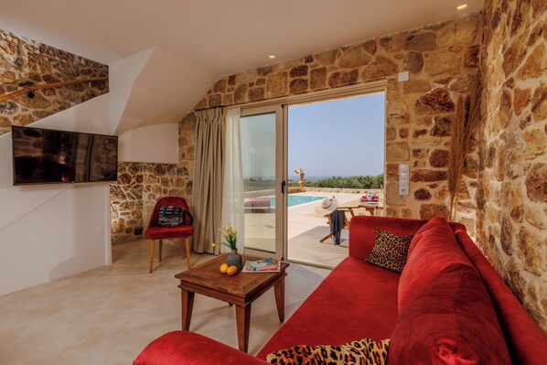 A living room with a stone wall, a red sofa, a wooden coffee table, and a TV. - Minoan Villas (Malia)