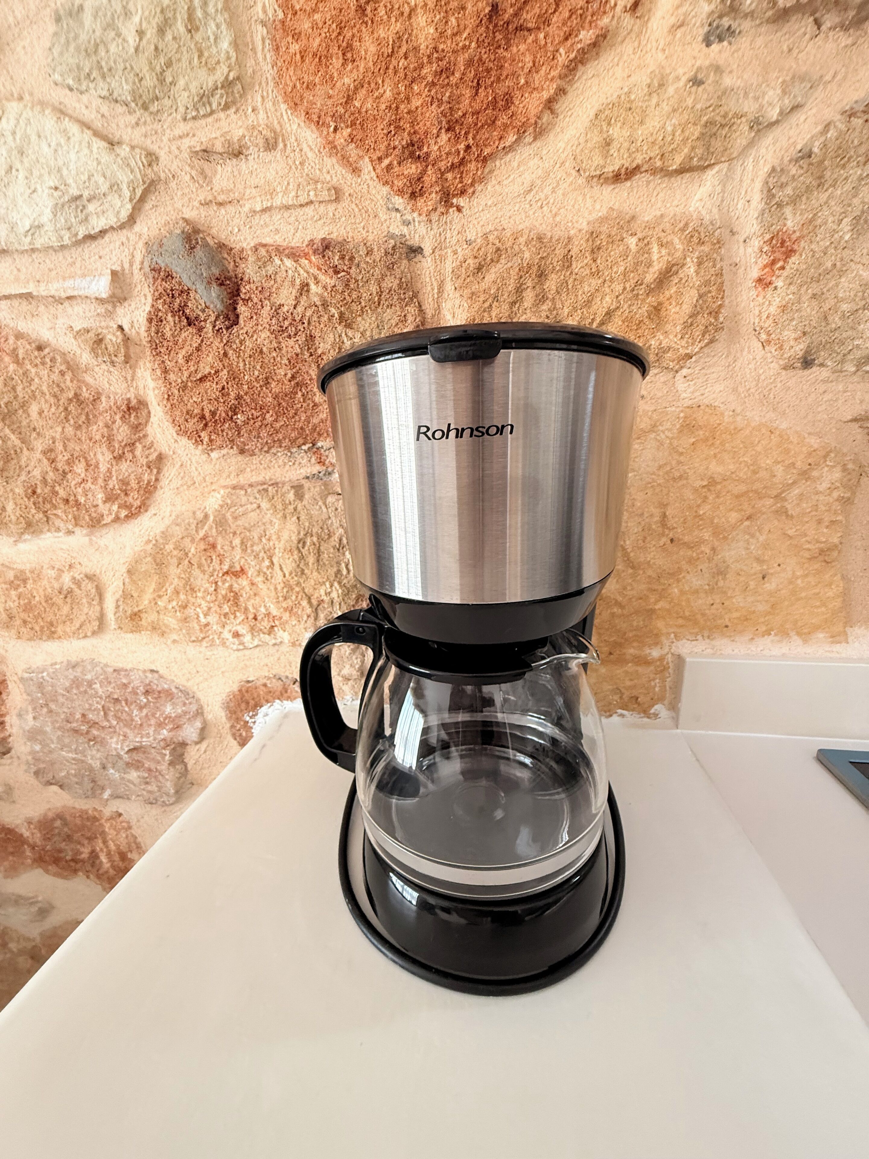 A stainless steel coffee maker with a clear carafe, placed on a white countertop against a stone wall.