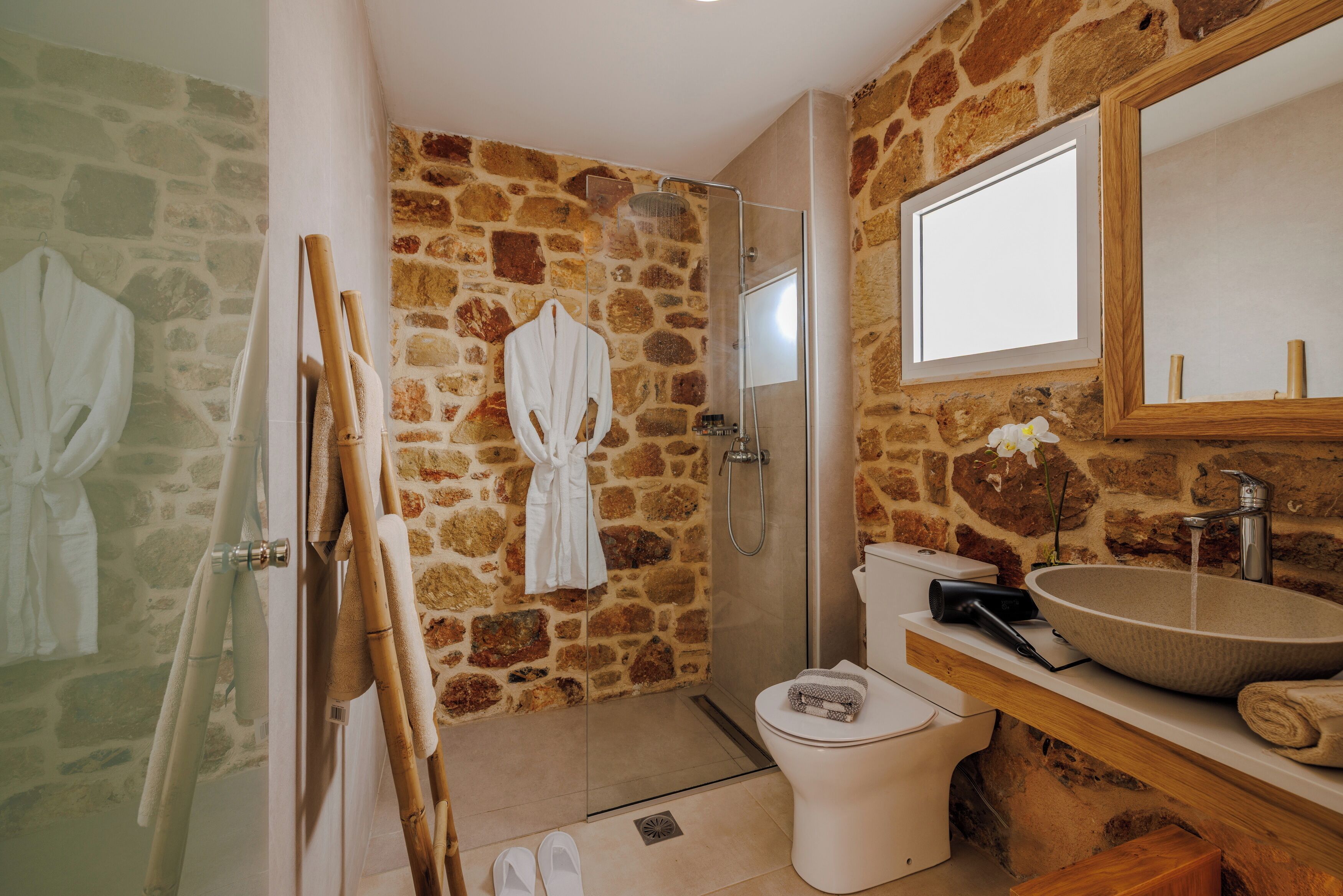 A bathroom with a stone wall, a glass-enclosed shower, a white toilet, and a wooden vanity with a large sink.