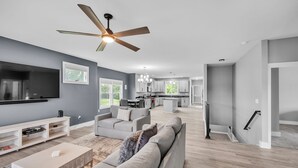A modern living room with a large sofa, a coffee table, and a TV. There is a ceiling fan, a kitchen in the background, and a staircase. - Pet-friendly Getaway With Golf & Private Patio Retreat 4 Bedroom Home (South Bend)