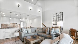Una sala de estar moderna con un sofá gris, una mesa de centro de madera y un sillón blanco. Cuenta con una zona de cocina con gabinetes blancos y un salpicadero de ladrillo. Un espejo grande cuelga en la pared, y se ve una escalera al fondo.
