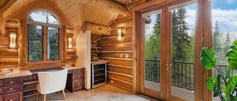 A log cabin interior with a wooden desk, chair, and large windows offering a view of the forest.