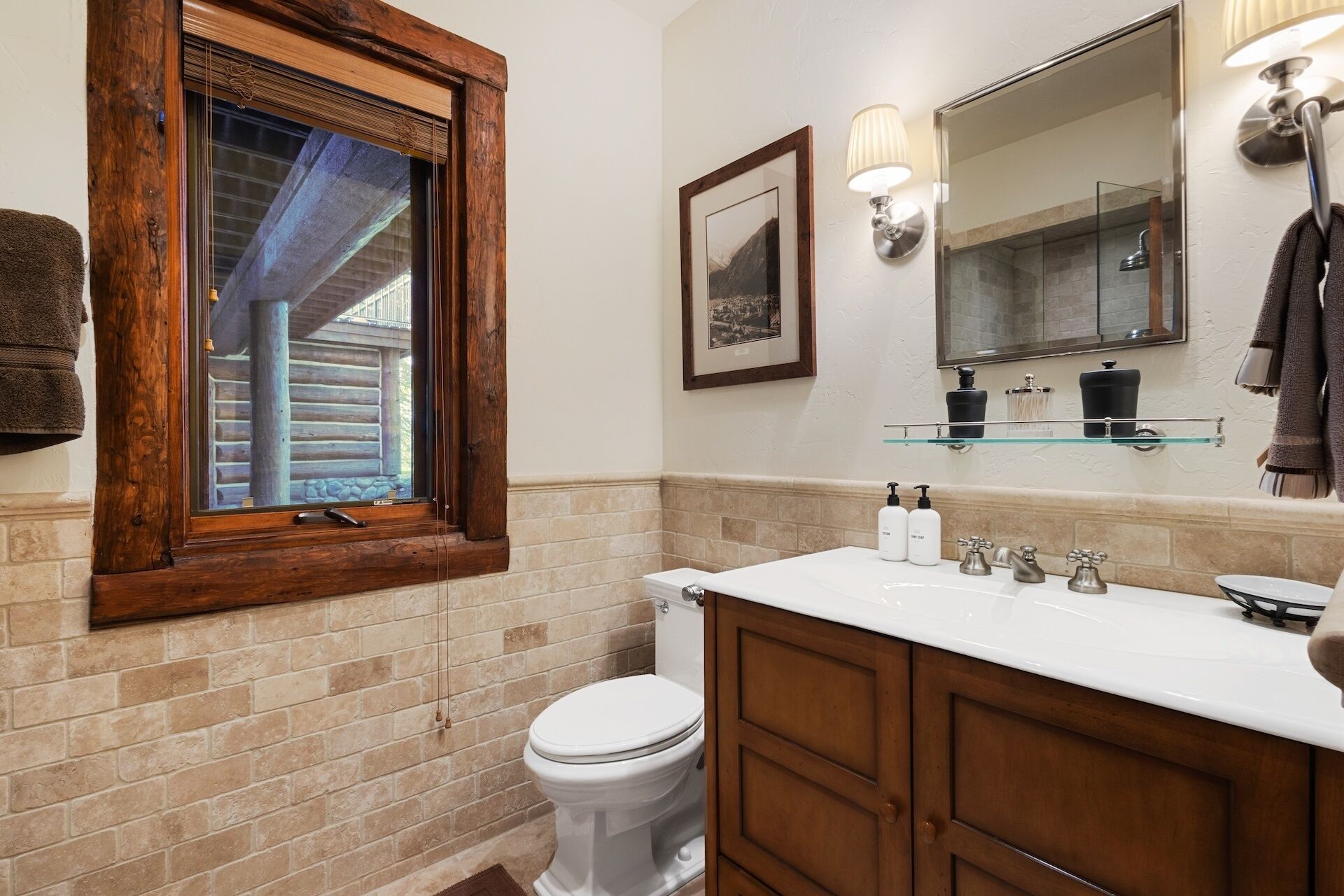 A bathroom with a wooden-framed window, a white toilet, a white sink with a mirror above, and a towel rack with towels.