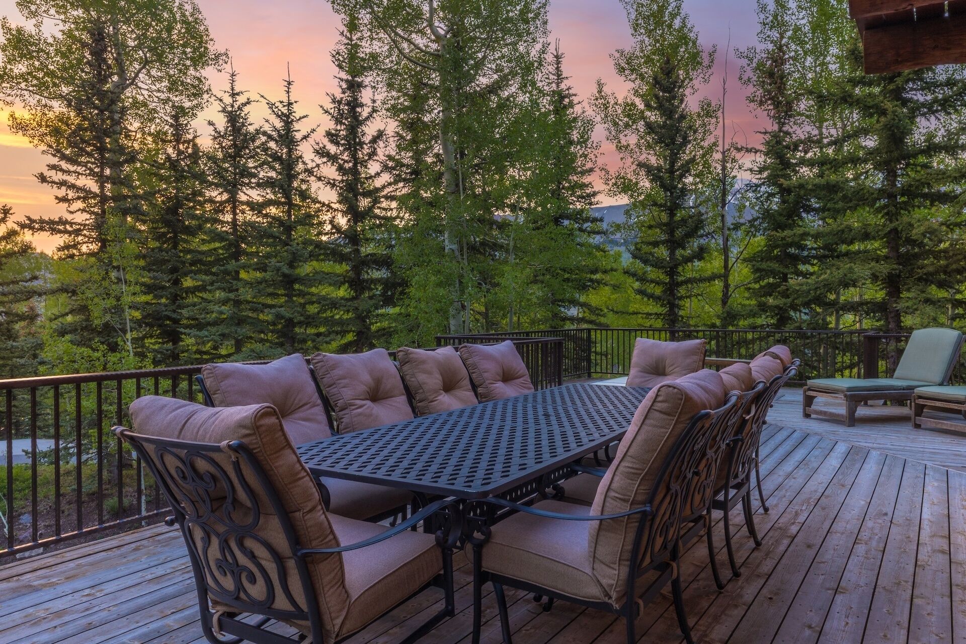 A wooden deck with a metal table and chairs, surrounded by tall trees and a sunset in the background.