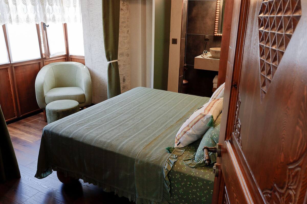 Una habitación con una cama, una silla, un taburete pequeño, una puerta de madera y una ventana con cortinas.