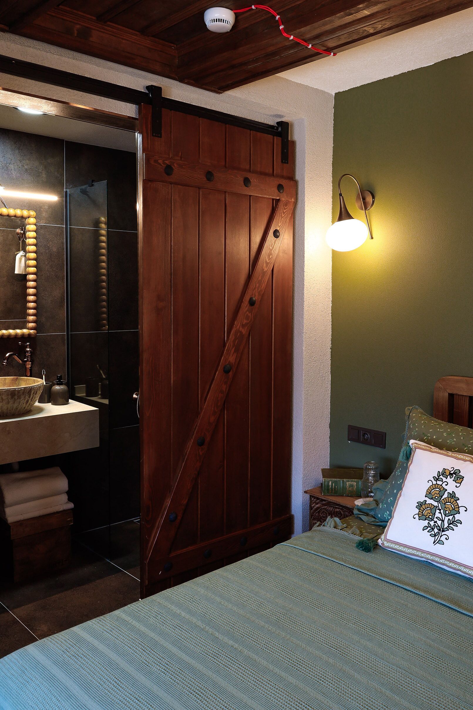 Una habitación con puerta corredera de madera, un lavamanos, una cama con colcha estampada y una lámpara de pared.