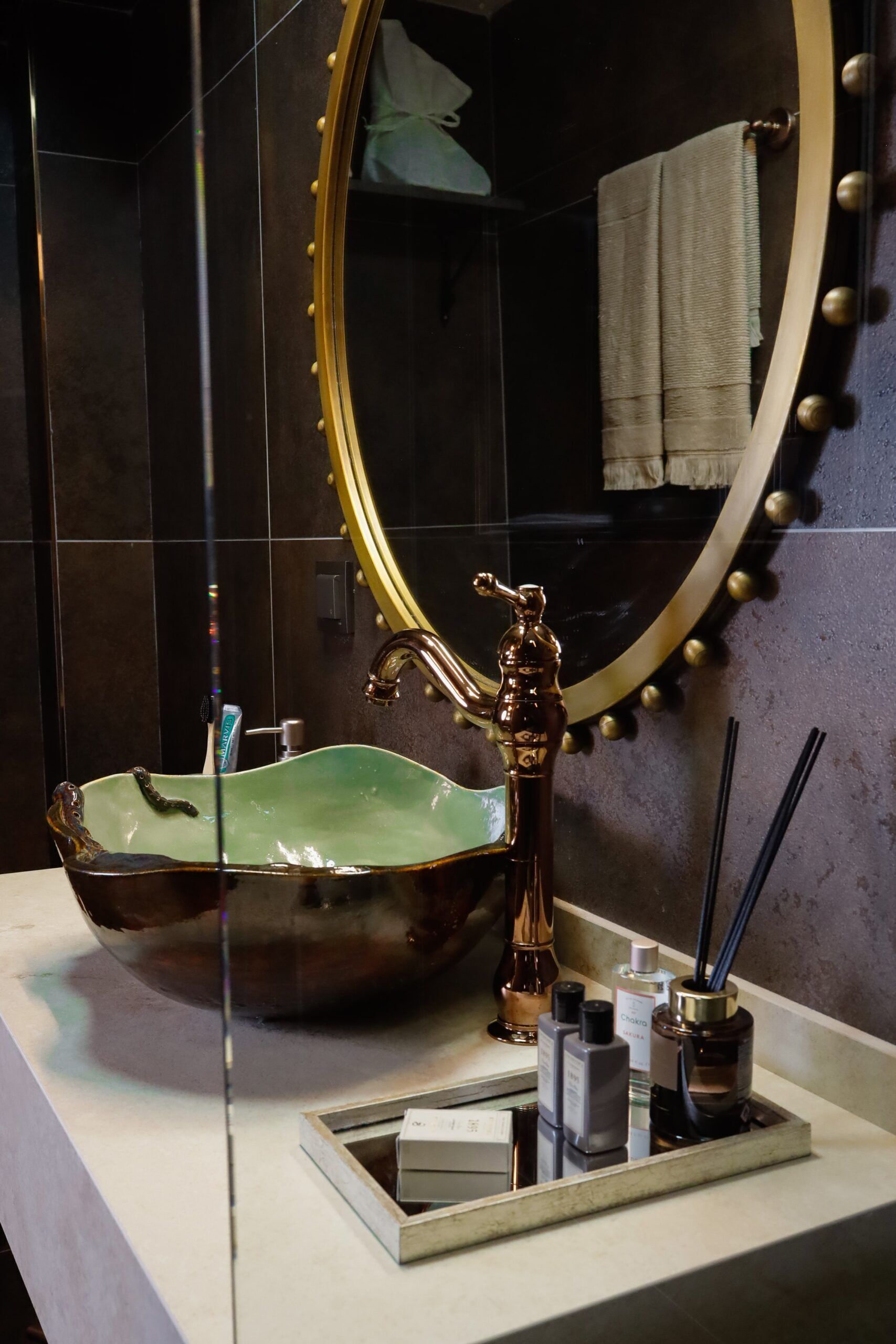 Un baño con un espejo ovalado grande, un lavabo de cerámica verde y un grifo dorado.