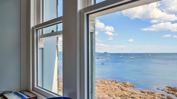 Quarto com grande janela vista para uma praia, barcos e céu claro.
