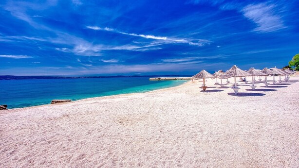 A beach with white sand and clear blue water. There are several thatched umbrellas and a few people in the distance.