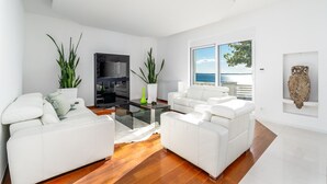 A modern living room with white sofas, a glass coffee table, and large windows offering a view of the sea.