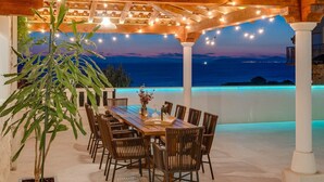 An outdoor dining area with a wooden pergola, string lights, and a view of the sea at dusk.