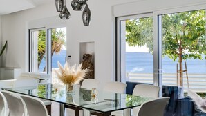 A modern dining area with a glass table, white chairs, and a view of the sea through large windows.