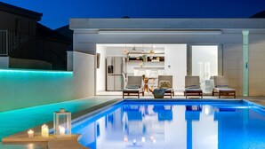 A modern outdoor pool area with a kitchen and seating area visible through an open door.