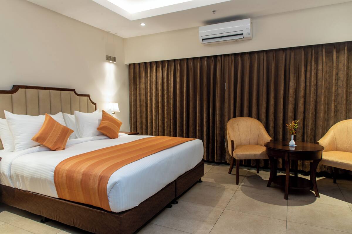 Quarto de hotel com uma cama grande, duas poltronas, uma mesinha e um fundo de cortina.