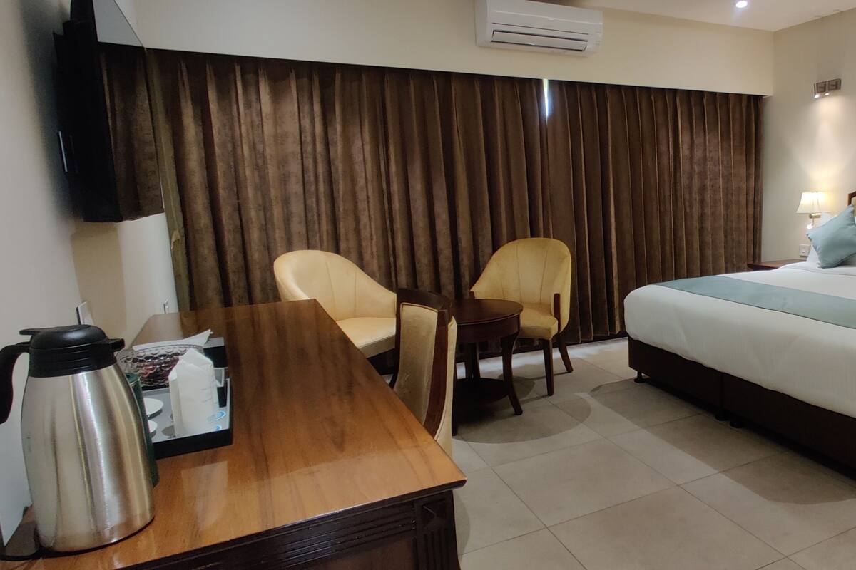 Quarto de hotel com cama, duas cadeiras, mesa de madeira com cafeteira, mesa de cabeceira com abajur e unidade de ar condicionado.
