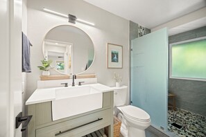 Ein modernes Badezimmer mit einem weißen Waschbecken, Spiegel und WC. Auf der Ablage steht eine kleine Pflanze und an der Wand hängt ein gerahmtes Bild.