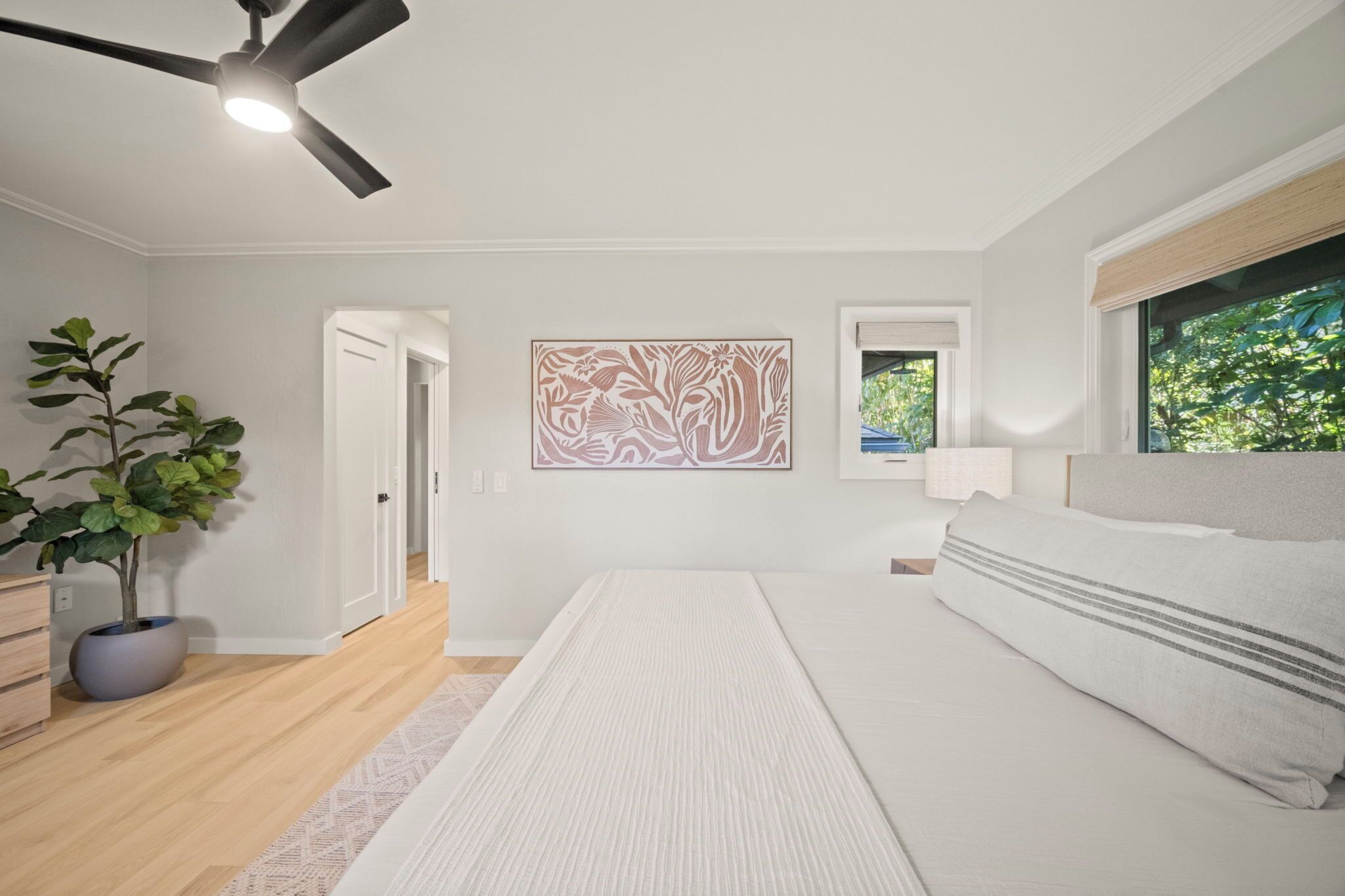 A bedroom with a bed, a ceiling fan, a potted plant, and artwork on the wall.