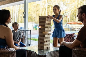 Eine Gruppe von Menschen spielt Jenga auf einem Tisch im Freien, mit Korbmöbeln und Blick auf Bäume sowie einen Basketballplatz im Hintergrund.