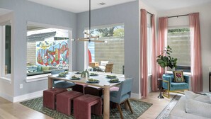 Un espace repas moderne doté d’une grande fenêtre, d’une table à manger dressée pour quatre personnes et d’un coin salon confortable avec un canapé et des poufs.