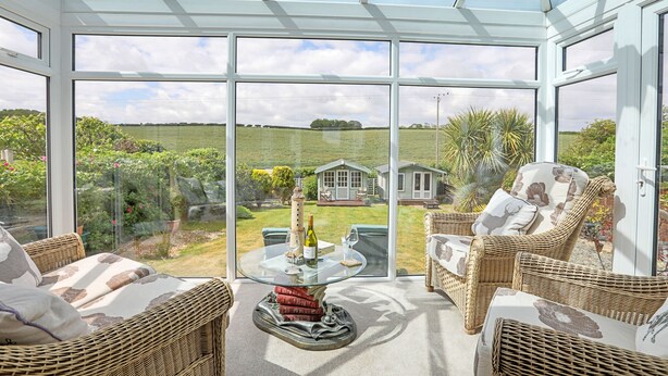 A conservatory with wicker furniture, a glass table, and a view of a house and garden. - Shoe Buttons Cottage at Gap Road (Filey)