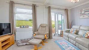 A living room with a sofa, armchair, TV stand, and a view of the outdoors through large windows. - Shoe Buttons Cottage at Gap Road (Filey)
