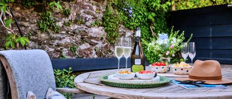 A round wooden table set outdoors with a bottle of wine, glasses, a hat, and a bowl of strawberries.