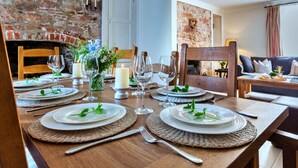 Un coin repas avec une table en bois dressée pour quatre personnes, entourée de chaises, avec un mur en briques et une cheminée en arrière-plan.