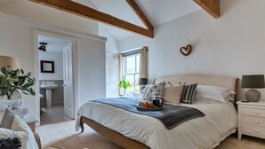 Une chambre avec un grand lit, des tables de chevet, une commode, une fenêtre avec des rideaux, une plante et une salle de bain visible au fond.