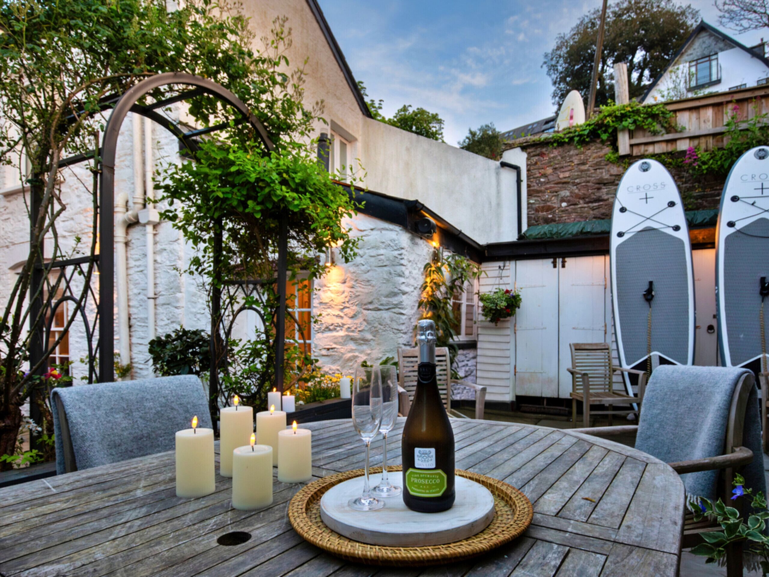 Een terras buiten met een houten tafel gedekt voor een maaltijd, omringd door kaarsen en een fles wijn.