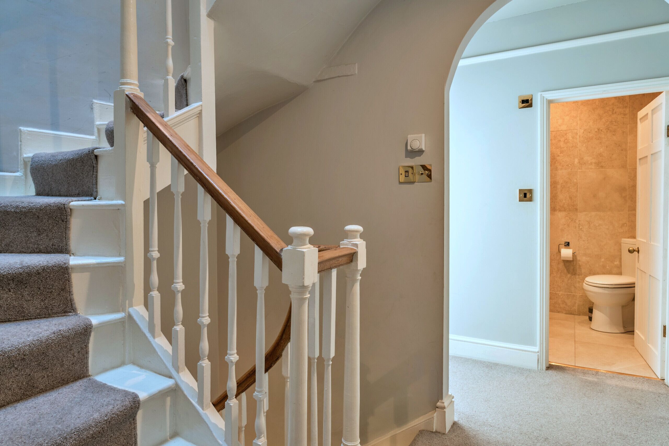 A staircase with white balusters and a wooden handrail leading to a bathroom with beige tiles and a white toilet.
