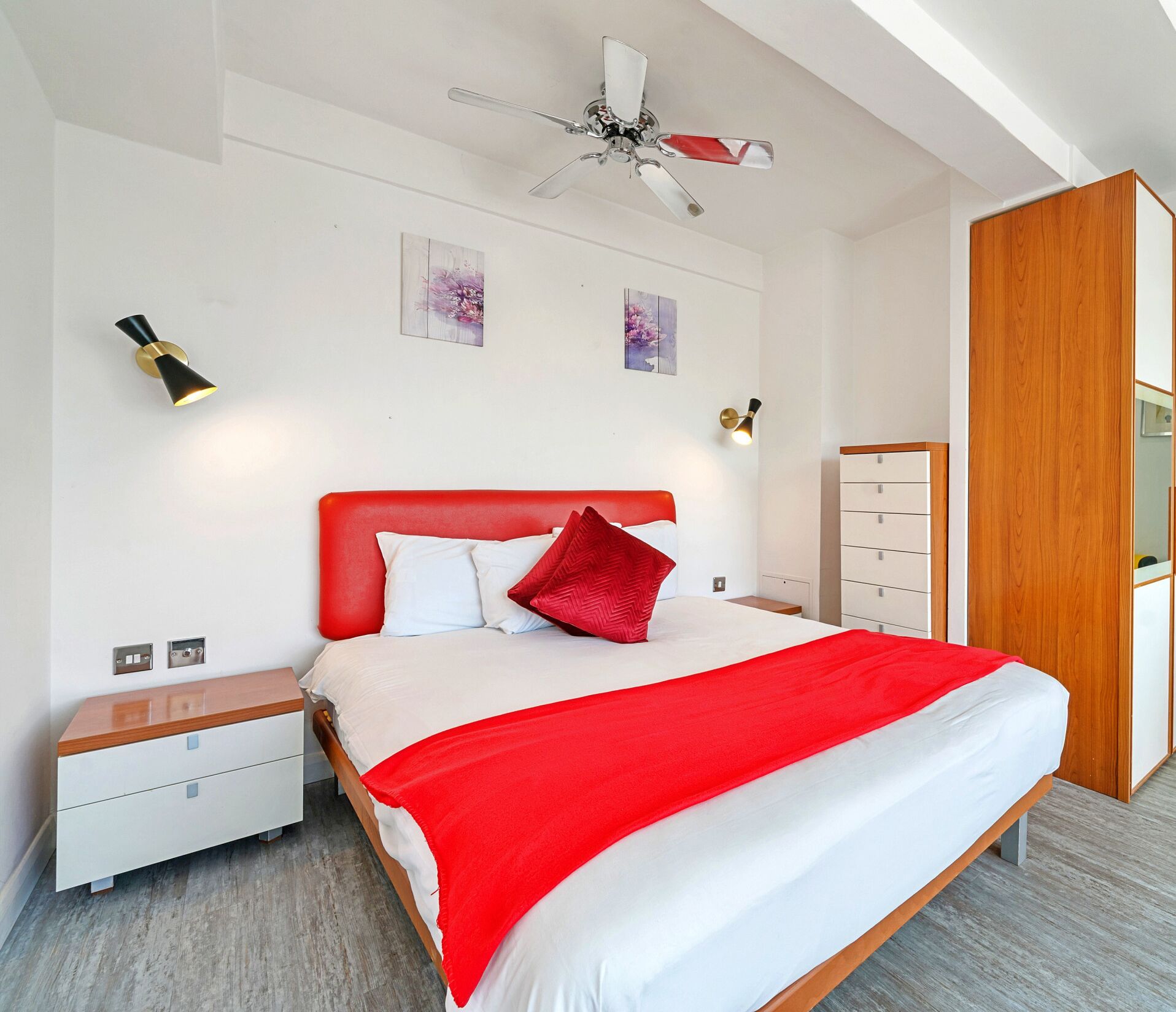 A modern bedroom with a red headboard, white bedding, a wooden nightstand, a dresser, and a ceiling fan.