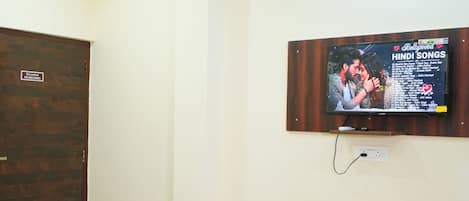 A hotel room with a bed, a television mounted on the wall displaying a Bollywood music channel, and a wooden door.