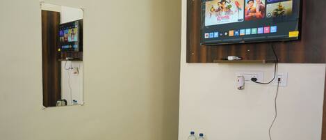 Habitación de hotel con televisión fija en la pared mostrando una película, una mesita con tetera, vasos y botellas de agua, y una cama con colcha estampada.