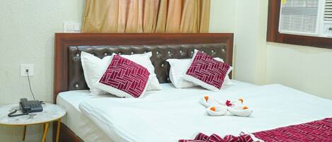 A neatly made bed with a white bedspread and two decorative pillows, a small bedside table with a phone, a brown rug, and a window with curtains.