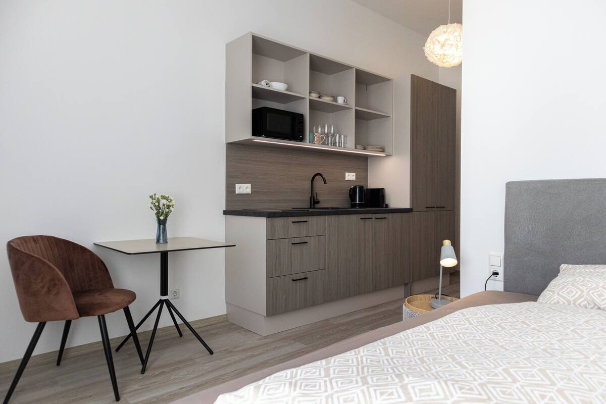 A modern kitchenette with a microwave, shelves, and a coffee maker. There is a small dining table with a chair, a bedside table with a lamp, and a bed with patterned bedding.