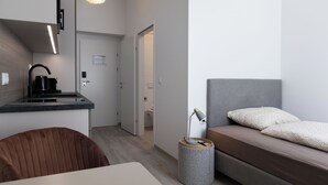 Una habitación moderna y funcional con kitchenette, una cama con almohadas estampadas y una mesita redonda con lámpara.