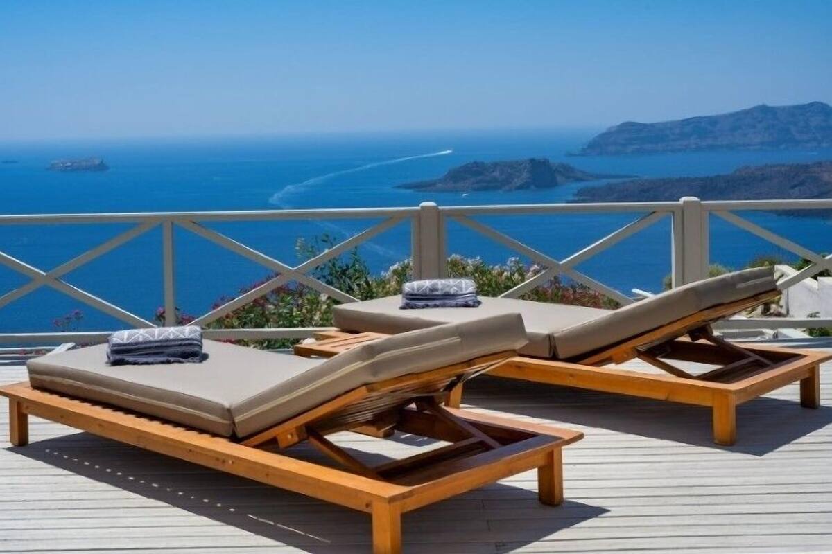 A wooden deck with two lounge chairs and a small table, overlooking a coastal view with clear blue waters and distant landforms.