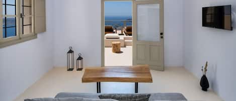 Una habitación con una mesa de madera, un sofá gris y vista al mar a través de una puerta abierta.