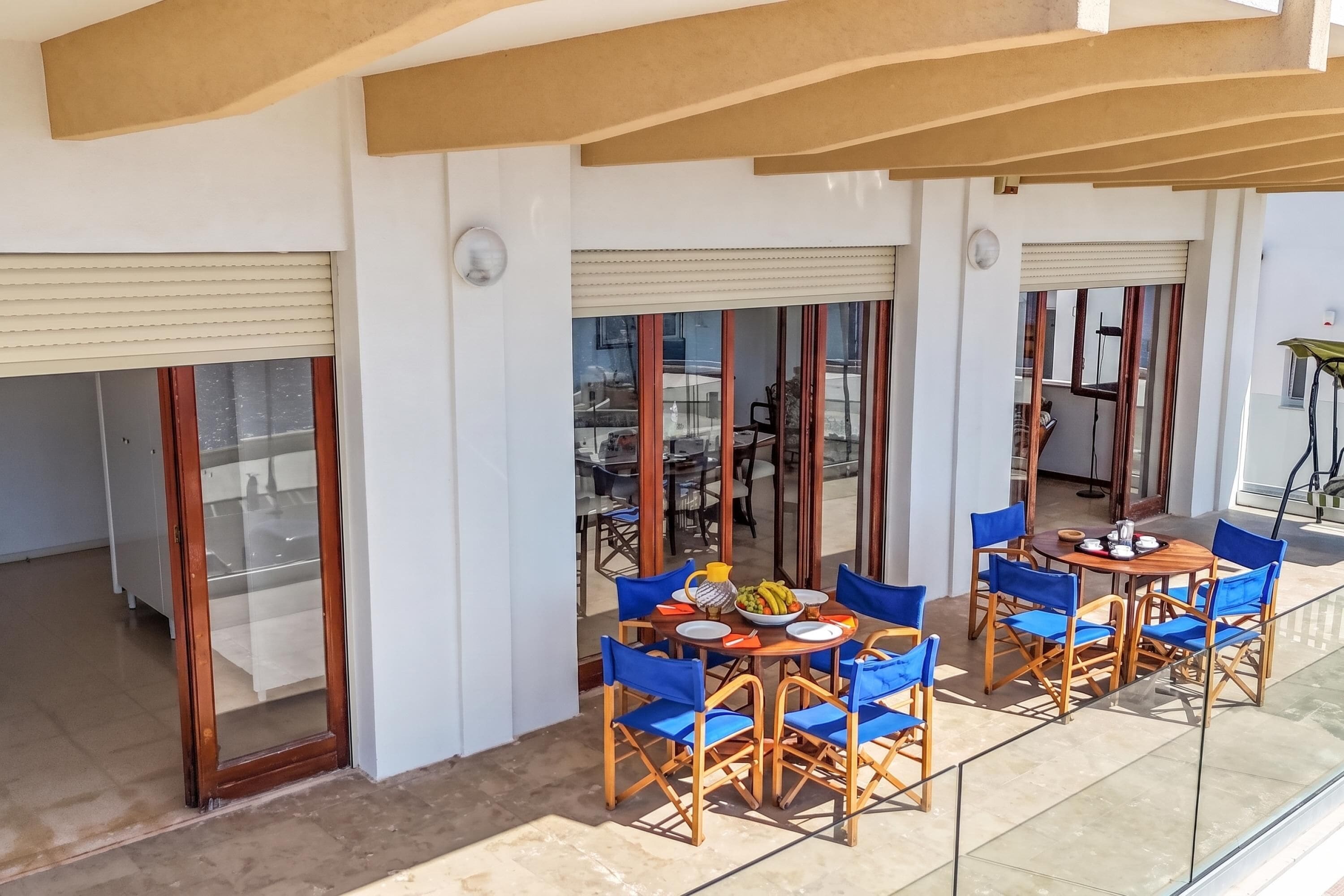 A covered outdoor seating area with tables and blue chairs, a glass partition, and a view of the street.