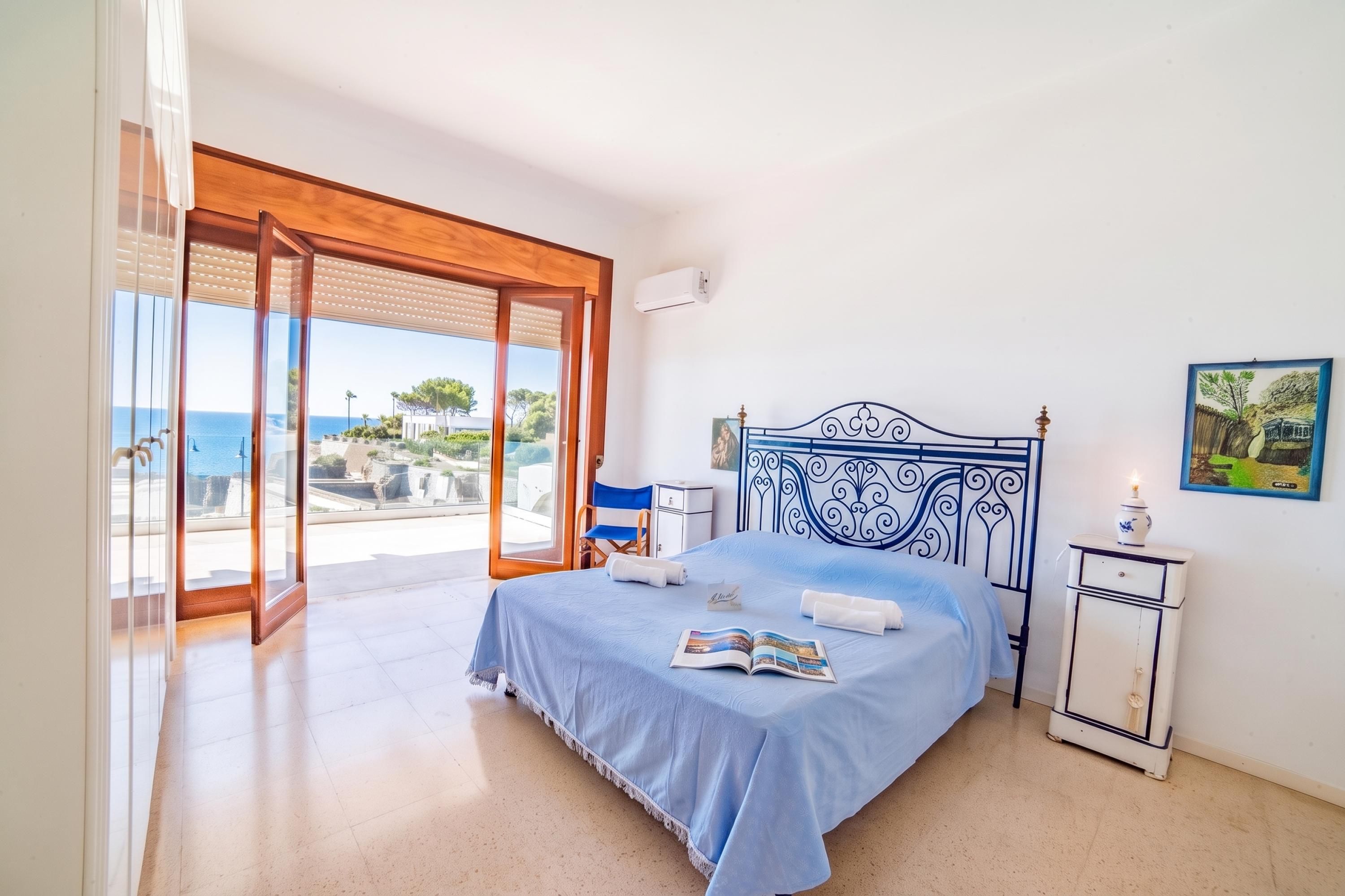 A bedroom with a bed, a nightstand, a window with a view of the sea, and a painting on the wall.