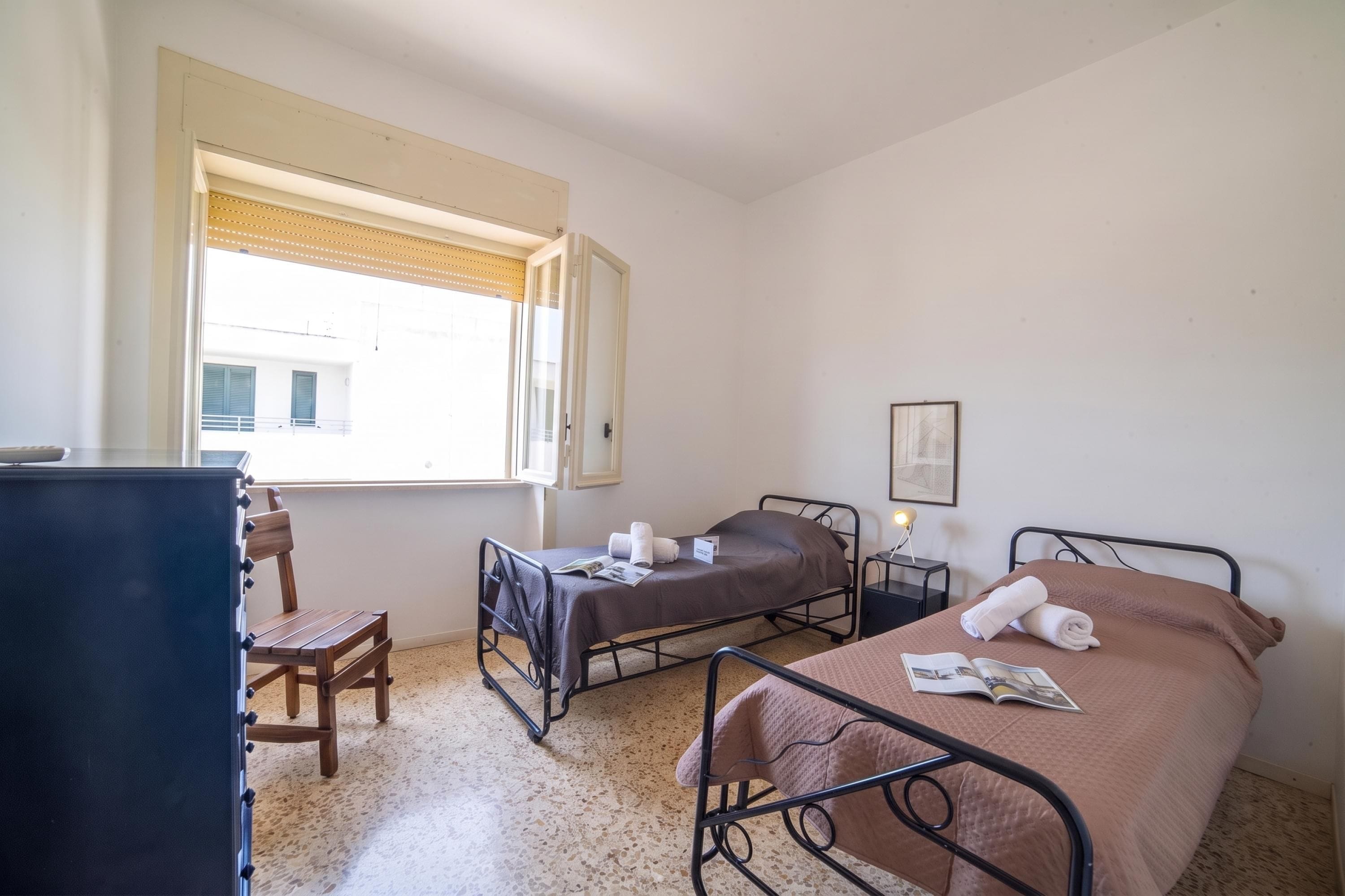A room with two beds, a chair, a desk, and a window with blinds.