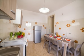 Uma cozinha moderna com área de refeições, equipada com geladeira, pia e mesa posta para dois, incluindo pratos, talheres e uma tigela com frutas.