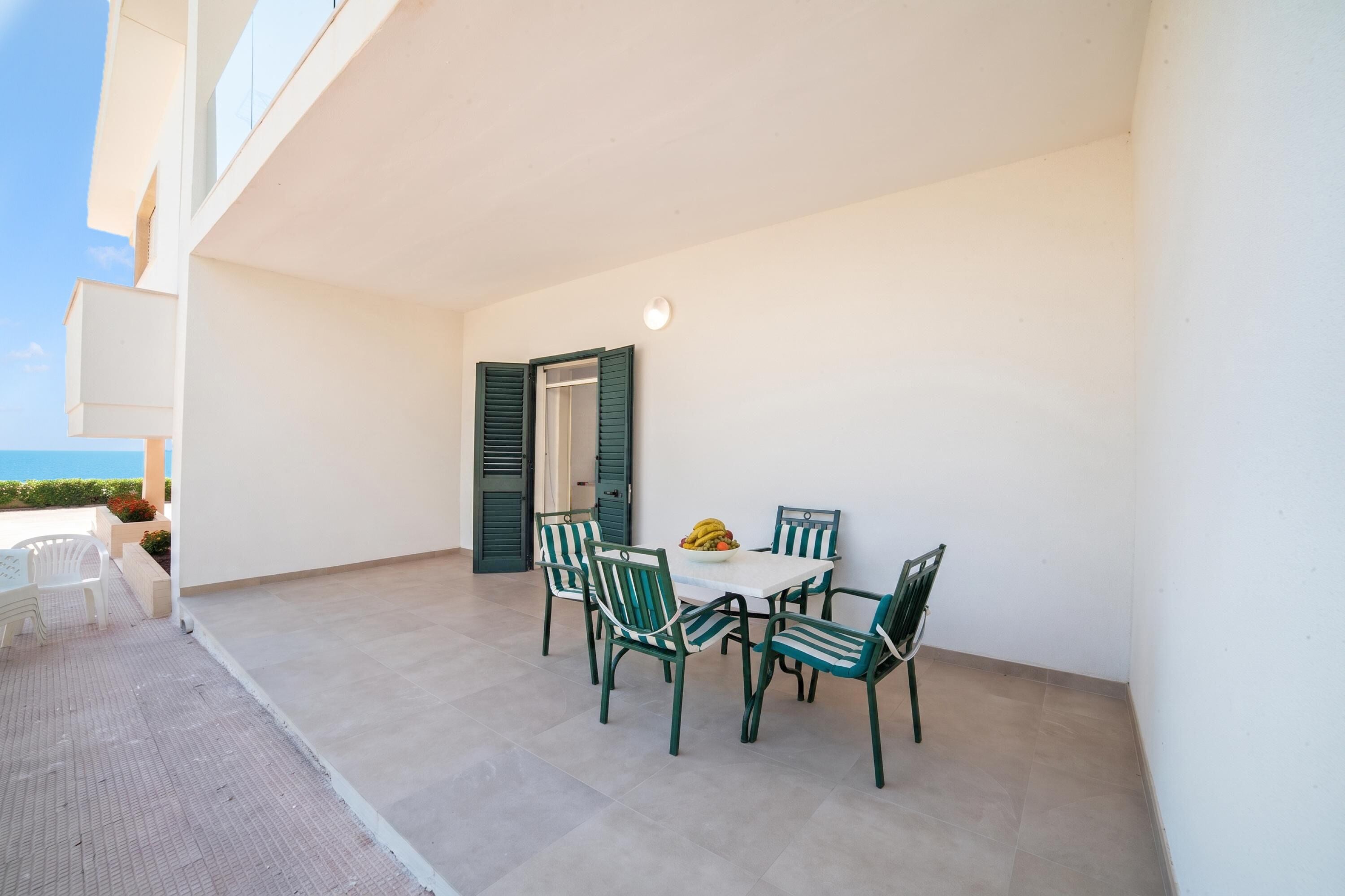 Um terraço ao ar livre com mesa e cadeiras, vista para o mar e um edifício com fachada branca.