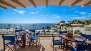 Terraço com mesa posta para refeições, vista para a costa com edifícios e árvores.