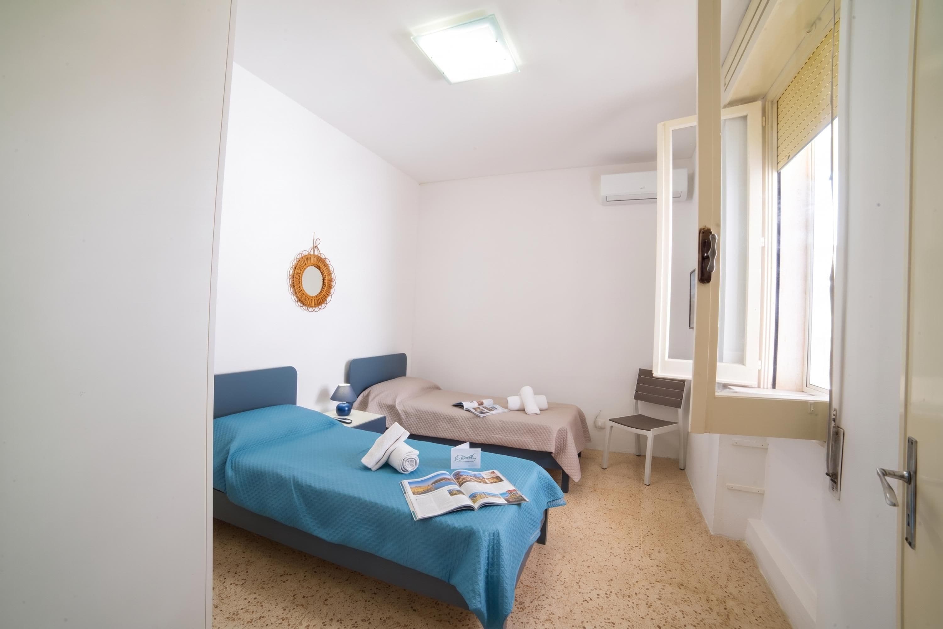 Une chambre d’hôtel avec deux lits, une petite table, une chaise, un climatiseur fixé au mur et une fenêtre avec des stores.