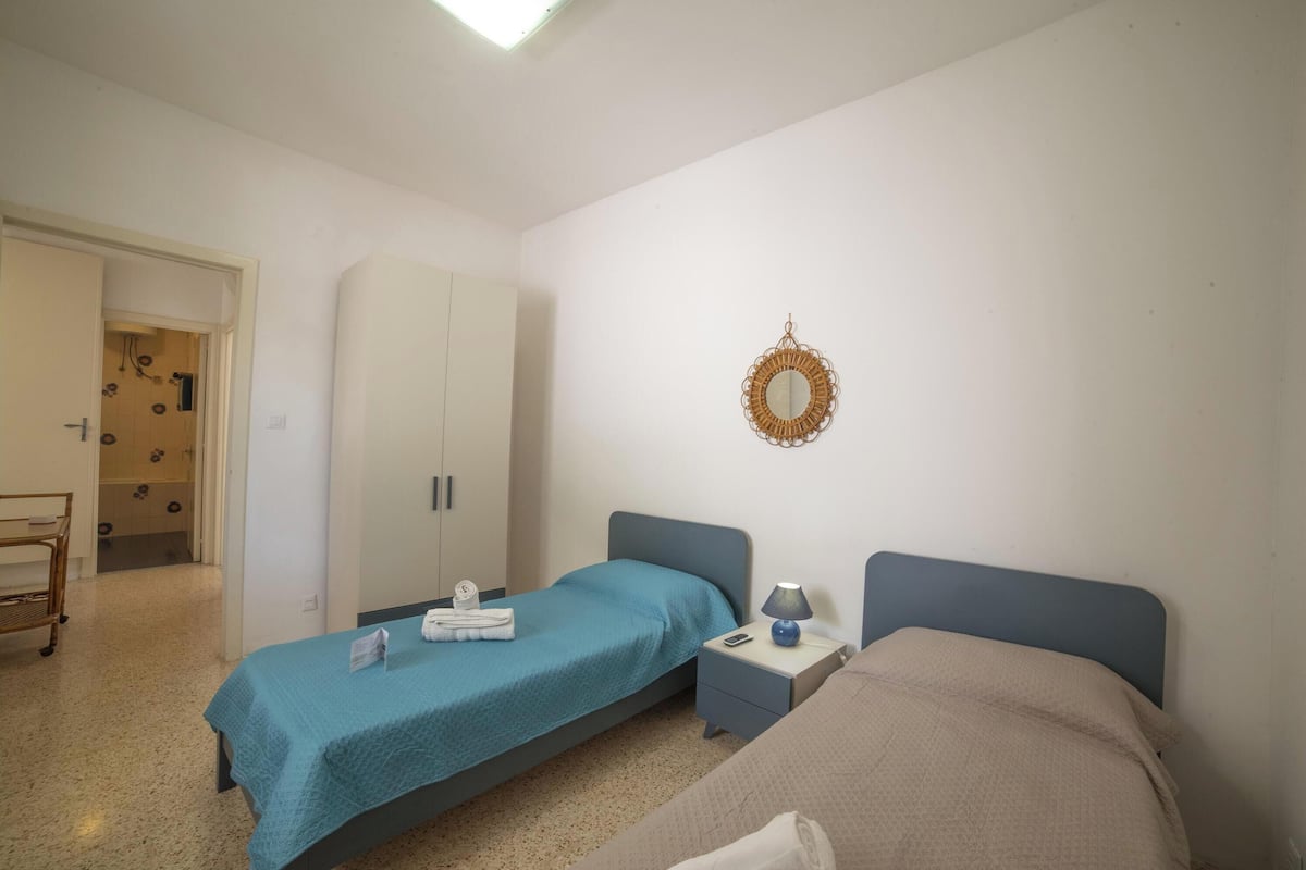 A bedroom with two beds, a wardrobe, a bedside table with a lamp, and a wall-mounted mirror.