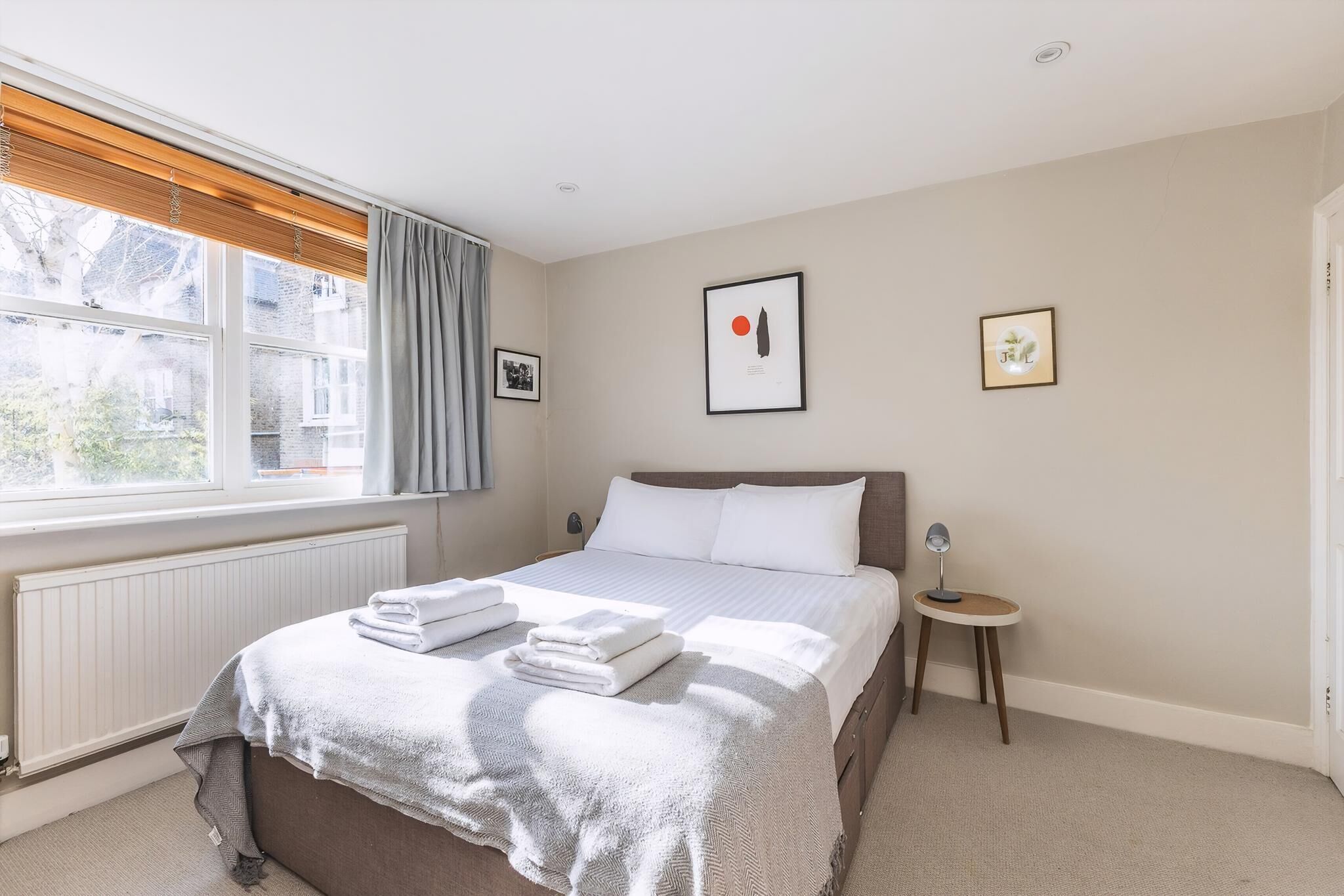 A neatly made bed with folded towels, a bedside table with a lamp, and a window with curtains.